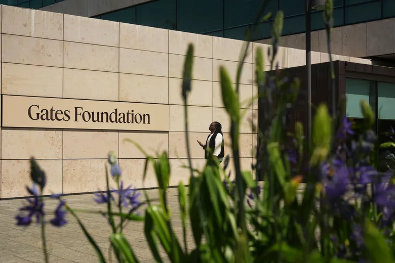 Hình minh hoạ bảng tên Gates Foundation trên tường đá toà nhà, một người đi bộ phía sau và hoa lá tím xanh tiền cảnh. Tin chính ngày 15/05/2026: Anthropic và Gates Foundation ký thỏa thuận 200 triệu USD đưa AI vào y tế, giáo dục và nông nghiệp.